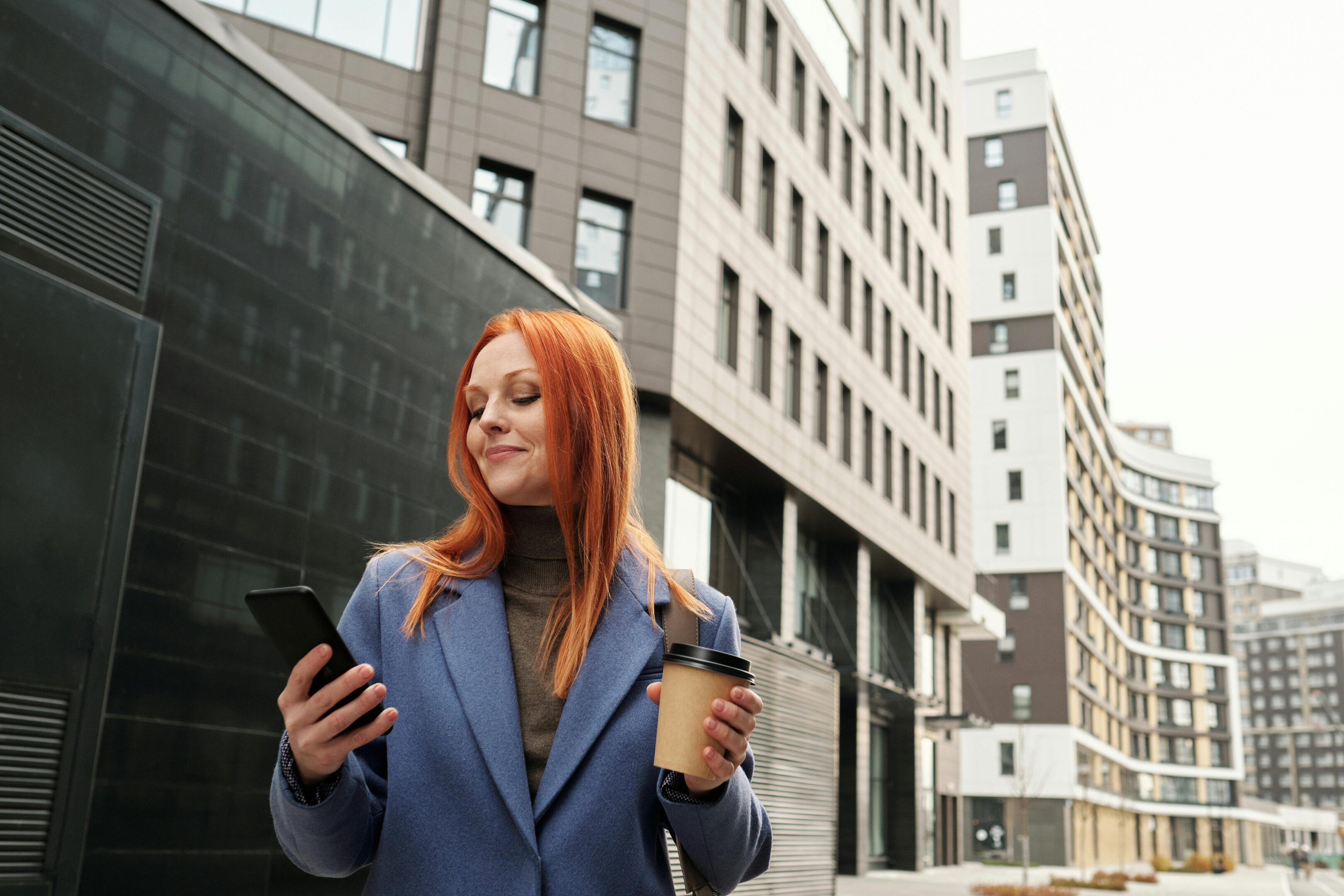 A woman with red hair holds a coffee cup while looking at her phone, appearing focused and engaged