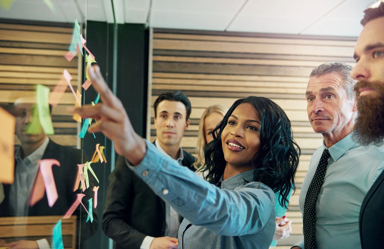 A group of diverse office workers collaboratively reviewing a board filled with colorful sticky notes.